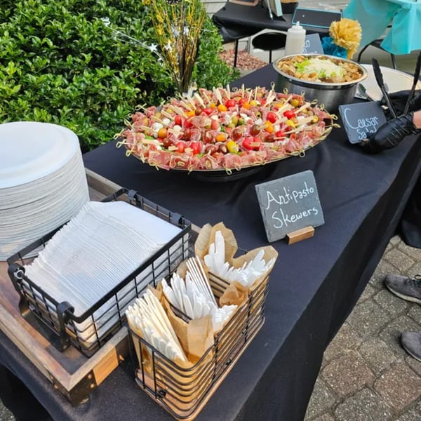 An outdoor buffet table with antipasto skewers, Caesar salad, plates, napkins, and cutlery.