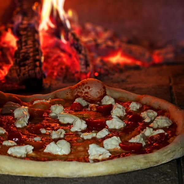 A pizza with tomato sauce, fresh mozzarella, and cherry tomatoes baking in a wood-fired oven.