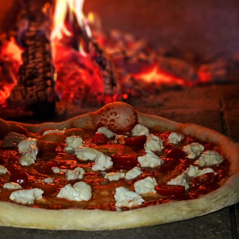 A pizza with tomato sauce, fresh mozzarella, and cherry tomatoes baking in a wood-fired oven.