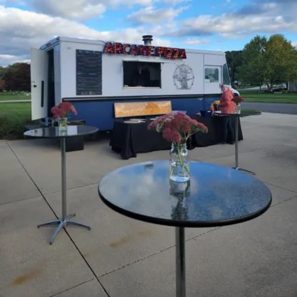 Arcane Pizza food truck with two tall tables and a black-draped serving table outside.
