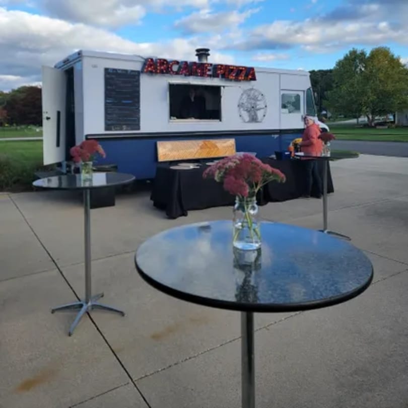 Arcane Pizza food truck with two tall tables and a black-draped serving table outside.