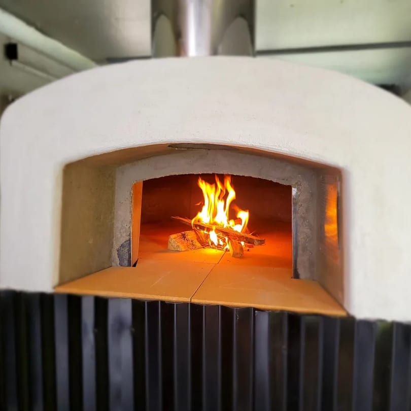 A white wood-fired pizza oven with a roaring fire inside, ready for cooking.