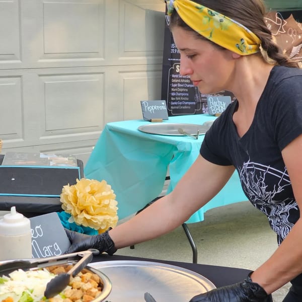 A woman in a yellow headband and black gloves prepares food at an outdoor event.