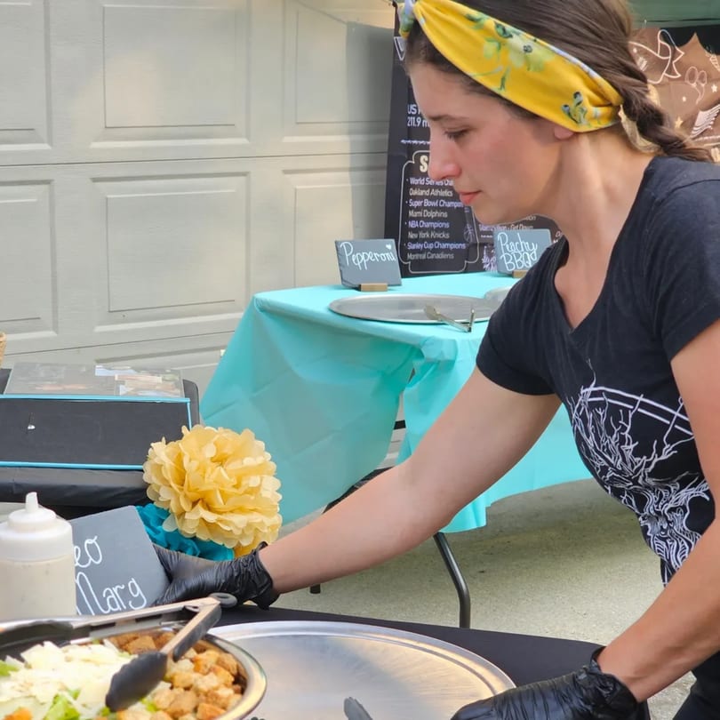 A woman in a yellow headband and black gloves prepares food at an outdoor event.