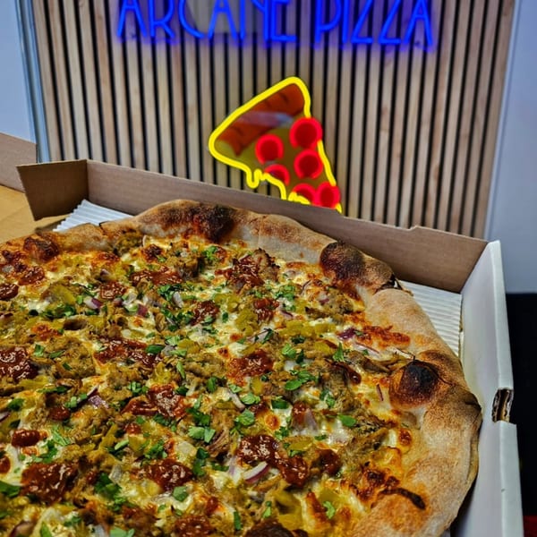A delicious pizza with various toppings in a box, with a neon Arcane Pizza sign in the background.