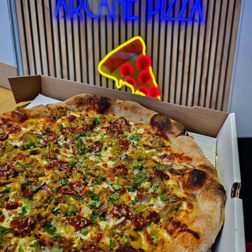 A delicious pizza with various toppings in a box, with a neon Arcane Pizza sign in the background.