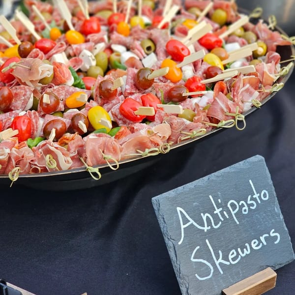 Antipasto skewers with meats, cheeses, olives, and tomatoes on a platter with a "Antipasto Skewers" sign.