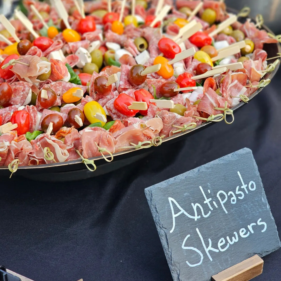 Antipasto skewers with meats, cheeses, olives, and tomatoes on a platter with a "Antipasto Skewers" sign.