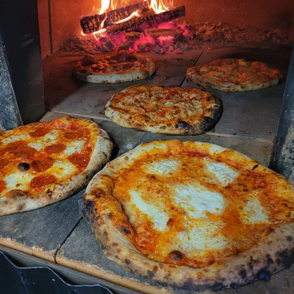Multiple pizzas baking inside a fiery wood-fired oven, with flames visible in the background.
