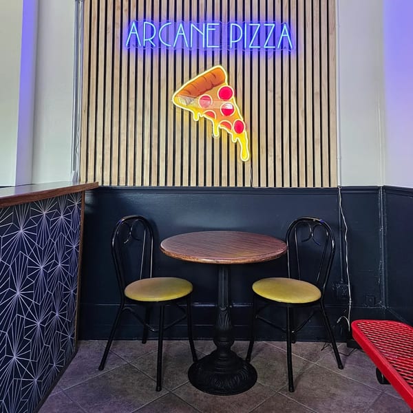 A small table with two chairs in a restaurant, with a neon "ARCANE PIZZA" sign and pizza slice.