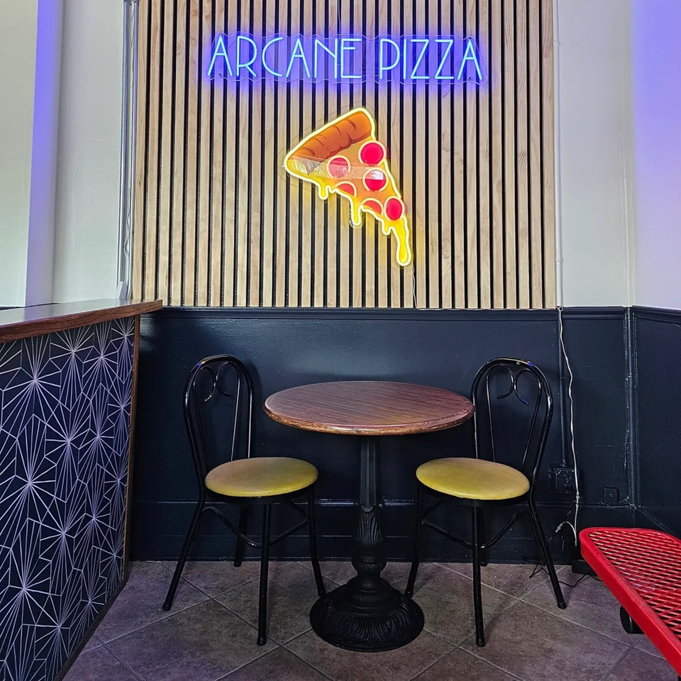 A small table with two chairs in a restaurant, with a neon "ARCANE PIZZA" sign and pizza slice.