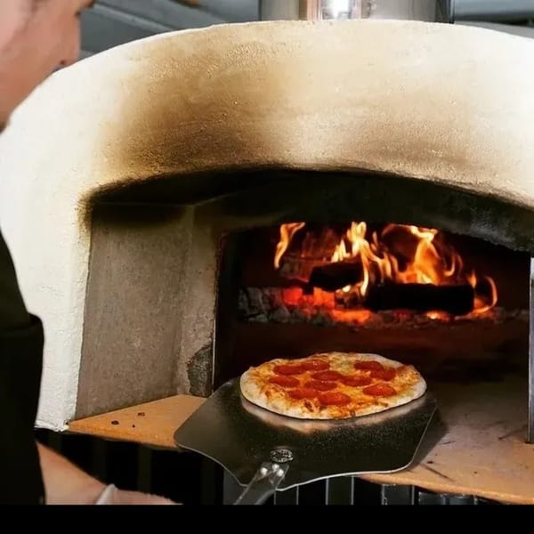 A chef slides a pepperoni pizza on a metal peel into a blazing wood-fired oven.