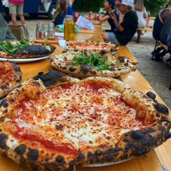Several wood-fired pizzas, some with arugula, on a long wooden table with people dining in the background.