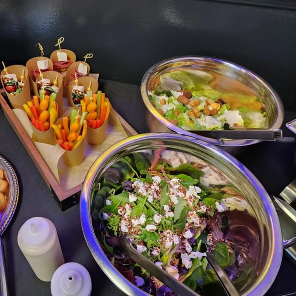 Buffet spread with two large salad bowls, vegetable cups, and charcuterie skewers on a dark table.