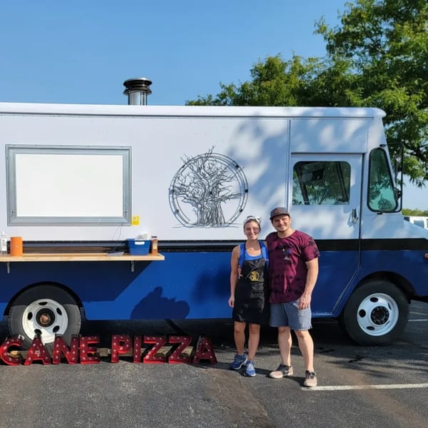 Two people stand proudly in front of their Cane Pizza food truck, featuring a tree logo.