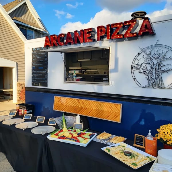 Arcane Pizza food truck with a buffet of fruit, cheese, and crackers on a black-draped table.