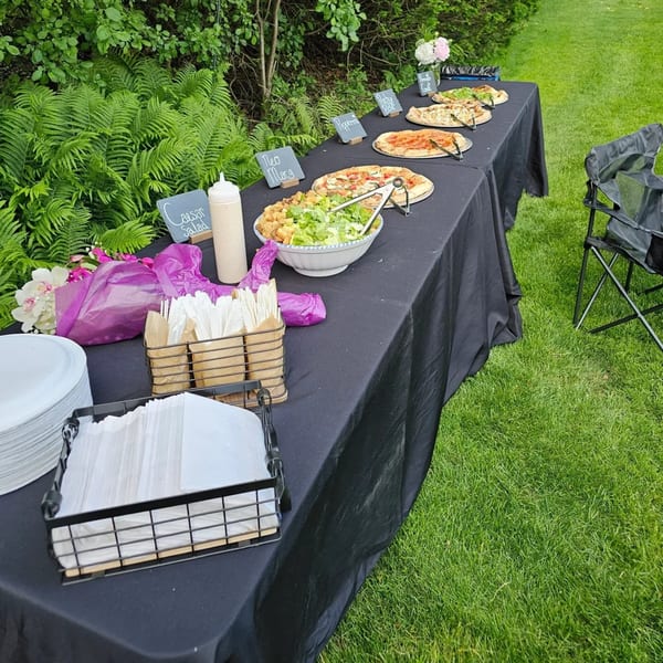 Outdoor buffet table with pizzas, Caesar salad, and serving supplies on a black tablecloth.