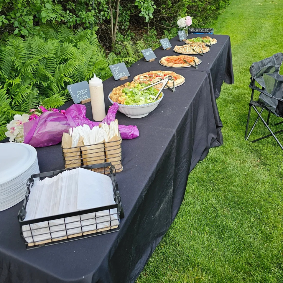 Outdoor buffet table with pizzas, Caesar salad, and serving supplies on a black tablecloth.
