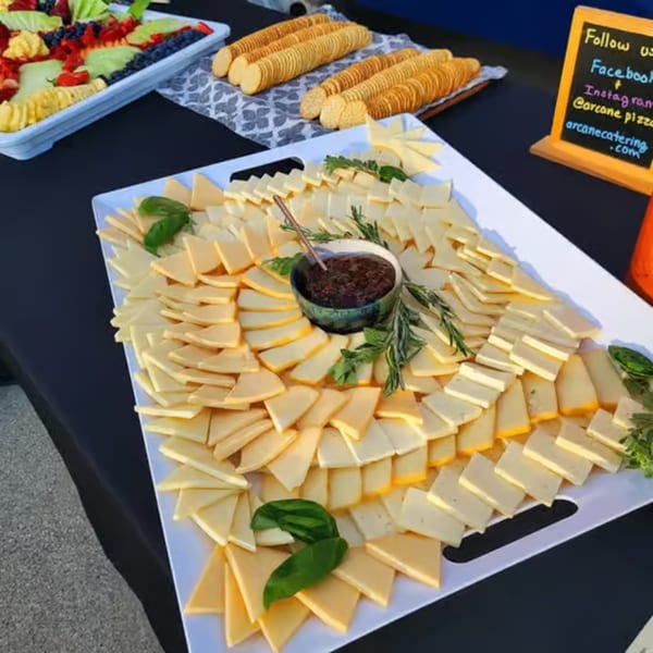 A cheese board with various cheeses, jam, and herbs, alongside crackers and a fruit platter.