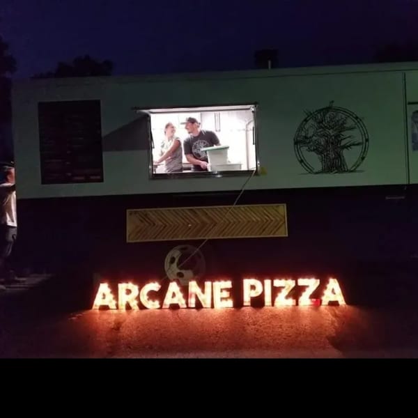 Arcane Pizza food truck at night with two people working inside and illuminated sign below.