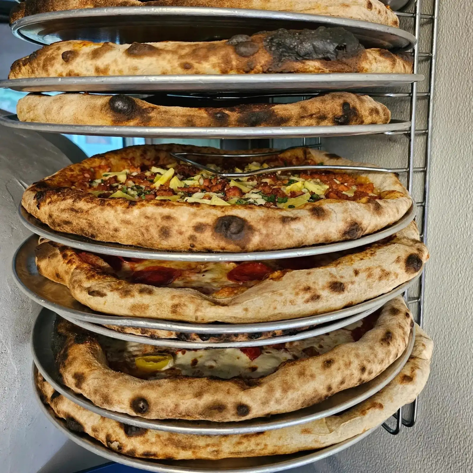 Multiple pizzas with crispy crusts and various toppings stacked on metal racks, ready to be served.