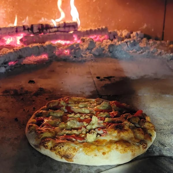 A pizza with chicken and red sauce cooking inside a wood-fired oven with glowing embers.